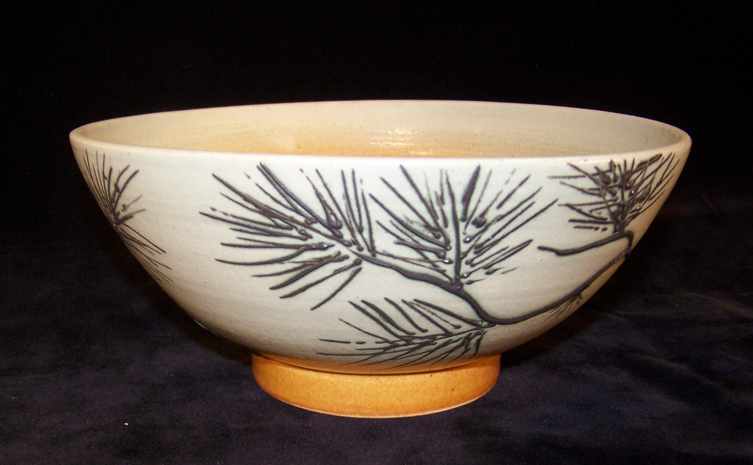 A wide shallow bowl that is likely thrown. It’s body is glazed white with dark brown pine branches and needles. It has a weighty foot that is glazed a muted orange.