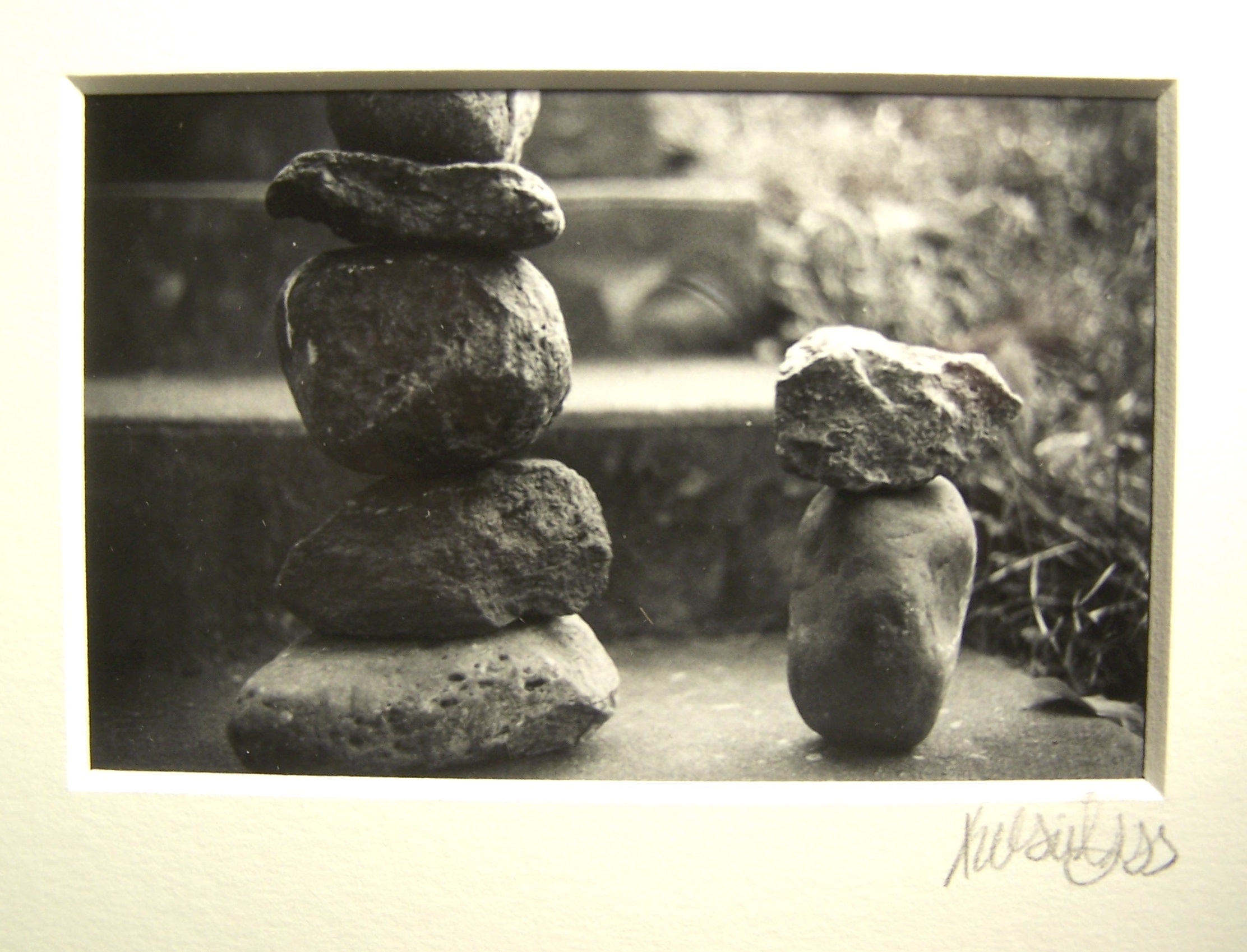 A black and white photograph of stones stacked on top of each other. On the left are five rocks of various sizes and shapes stacked and on the right is only two. Behind the stones is an out of focus staircase that ends behind the stack on the right creating a nice diagonal mirroring the two stacks. To the right of the staircase is some out of focus greenery.