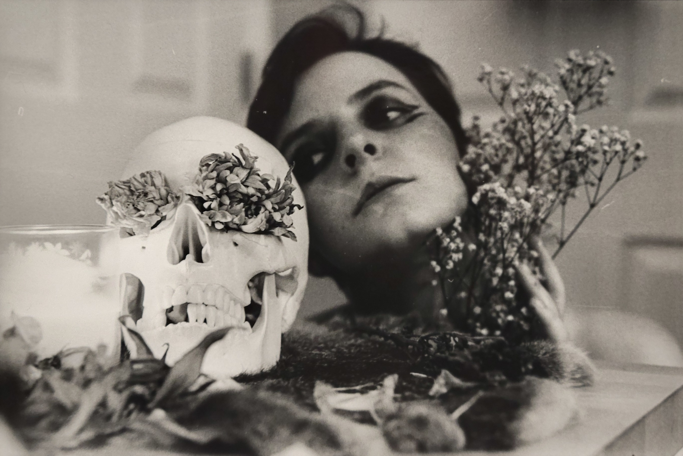 A still life black and white photo. A skull sits on a table, with flowers in its eyes, the skull sits next to a lit candle and other flowers. Next to the skull is the head of a person, who looks bodyless as they rest their head on the table. The figure is looking at the skull.