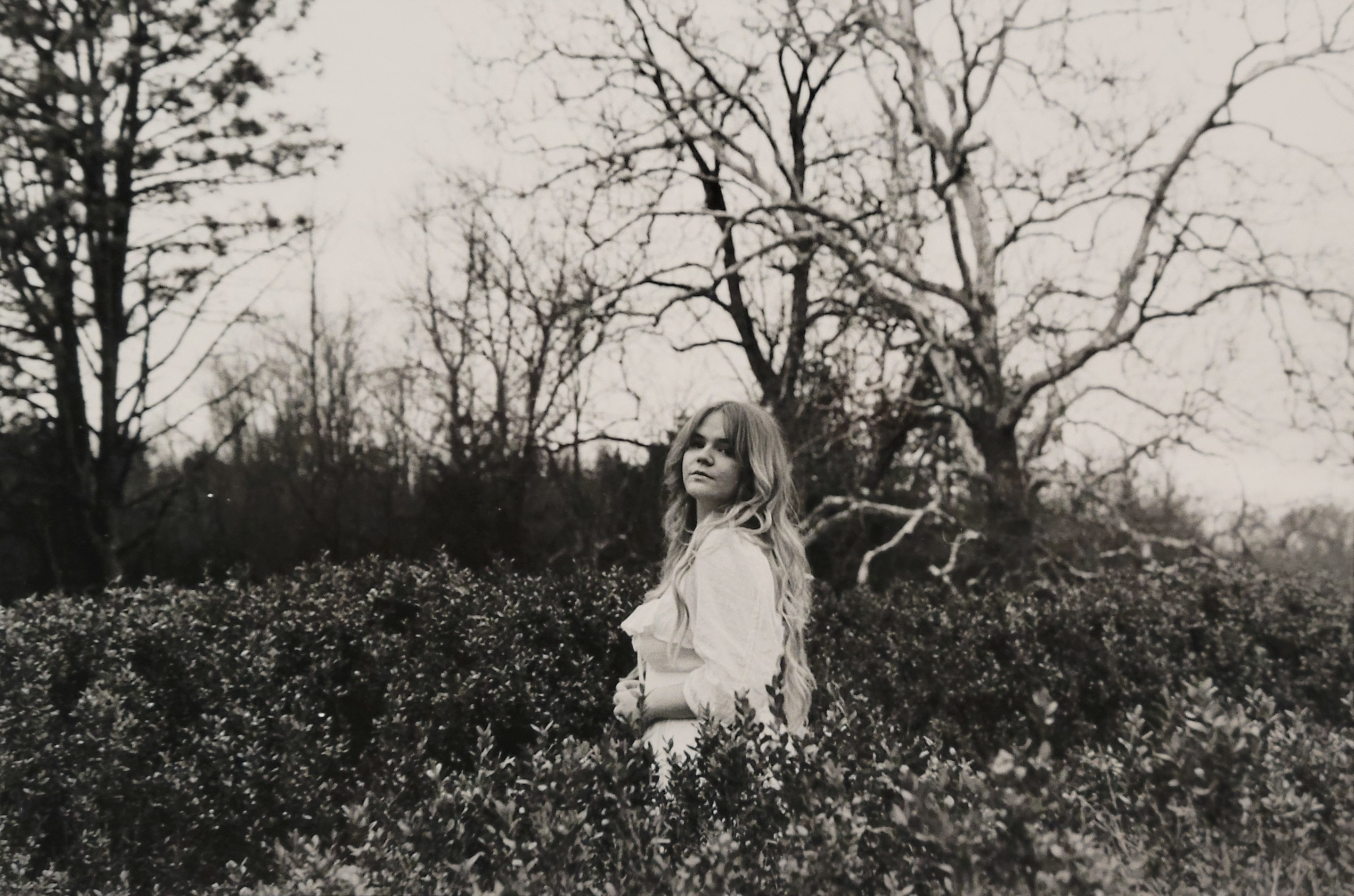 A landscape black and white photo. A woman stands in the center of a field of wildflowers, looking at the camera. She wears a simple white dress. Behind the figure to the left of the photo is a barren tree