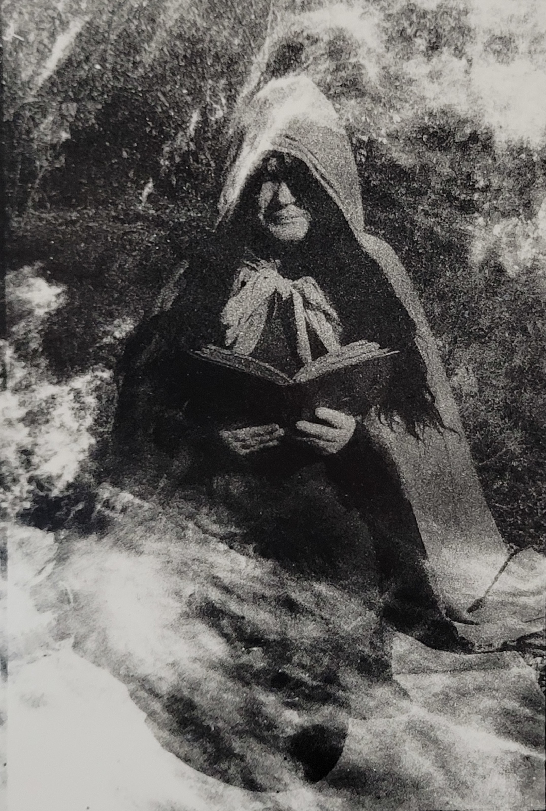 Black and White photograph of a figure, cloaked, kneels on the ground with a book in their hand ready to cast a spell. 