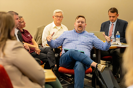 A man sitting in a chair is speaking and gesturing with his hands