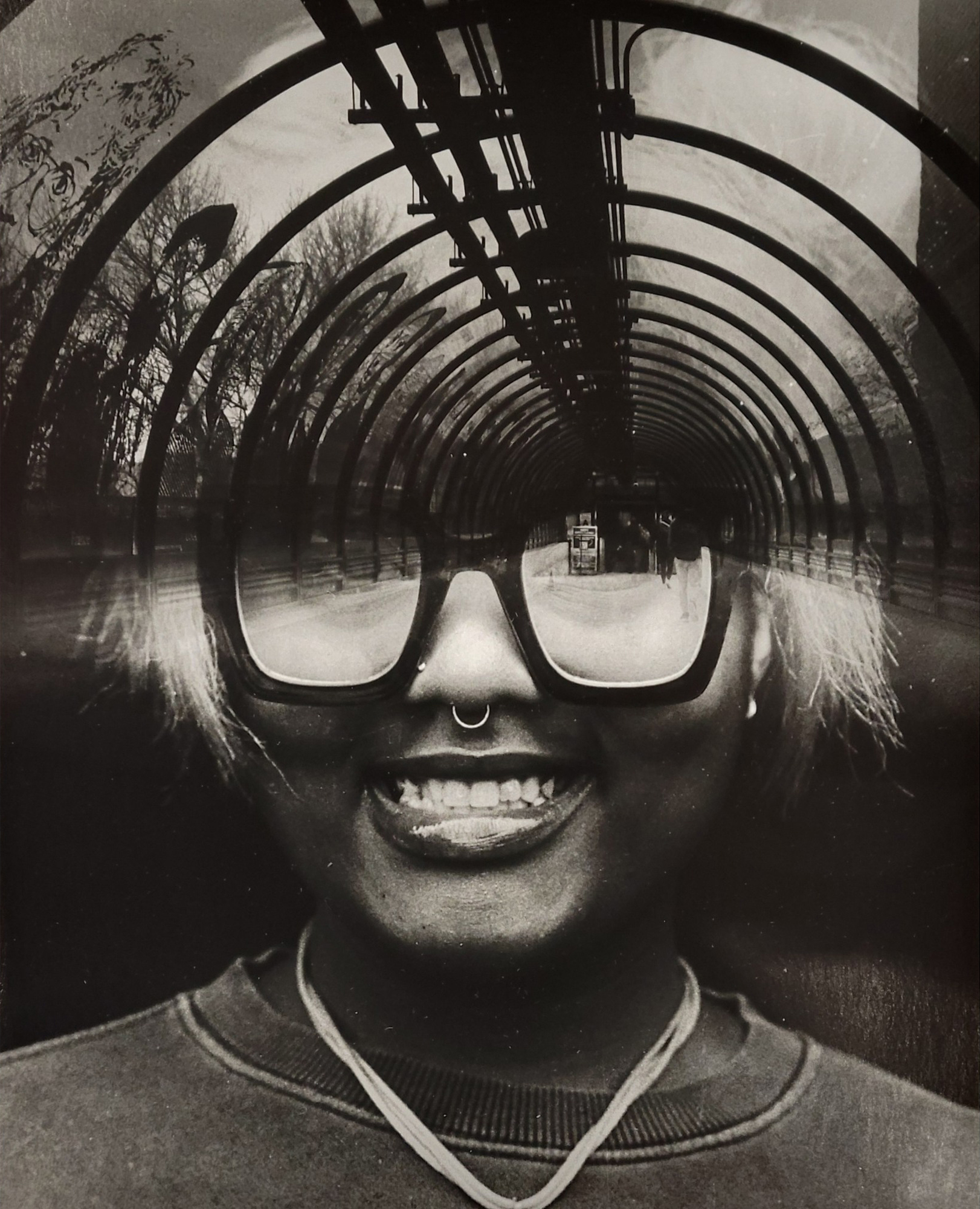 A black and white photograph of a girl smiling and looking directly at the camera. Viewed from the collarbone up, she has a nose ring and a septum piercing and is wearing a contrasting necklace creating a v in the lower part of the photo. The lower half of her face is clear and in focus, but the top of the photograph is layered on top with a view from Sinclair’s arched covered walkway featuring vinyl decals famous artists. Her thick glasses rounding down is a reverse repetition of the arched repeating structure that is moving to a one point perspective, focused just above her right eye.