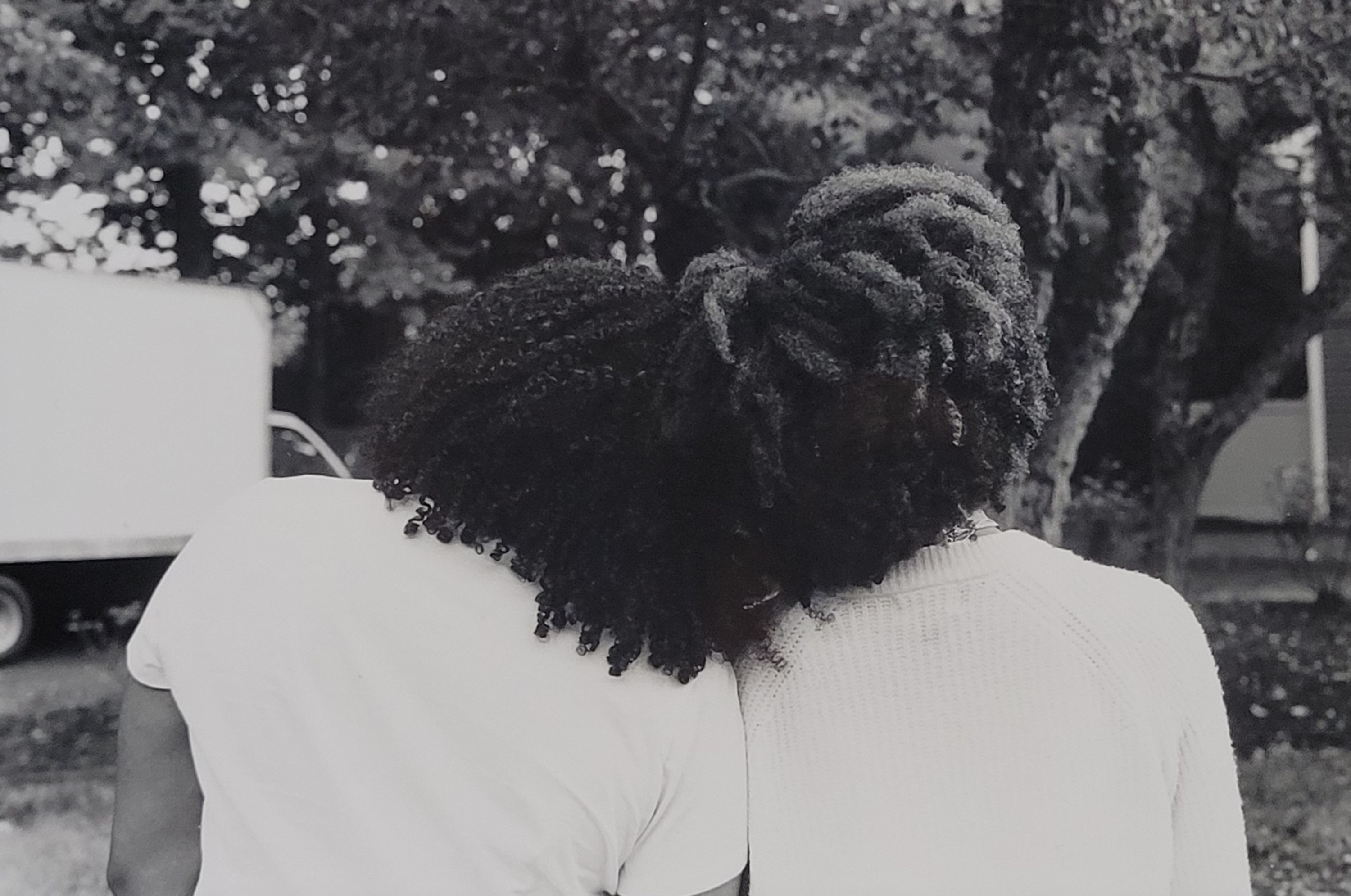 A black and white photograph of an image of two figures with foliage behind them. The figures are facing away from the camera, so that their natural curly hair is the focal point. They rest their heads against each other so that their hair blends together. Their dark hair contrasts with their white shirts, but blends with the leaves of the trees in the background.